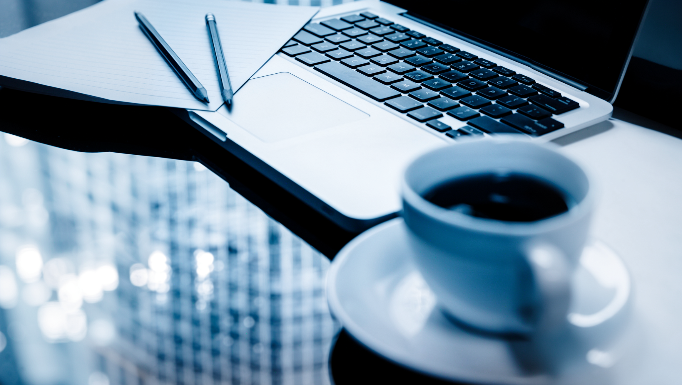 A close-up, shallow depth of field shot shows a laptop keyboard with black keys. To the left of the keyboard, two pencils rest on a piece of paper. In the foreground, a white coffee cup and saucer are visible, with a dark liquid inside. The background is blurred and bright, suggesting a well-lit environment.